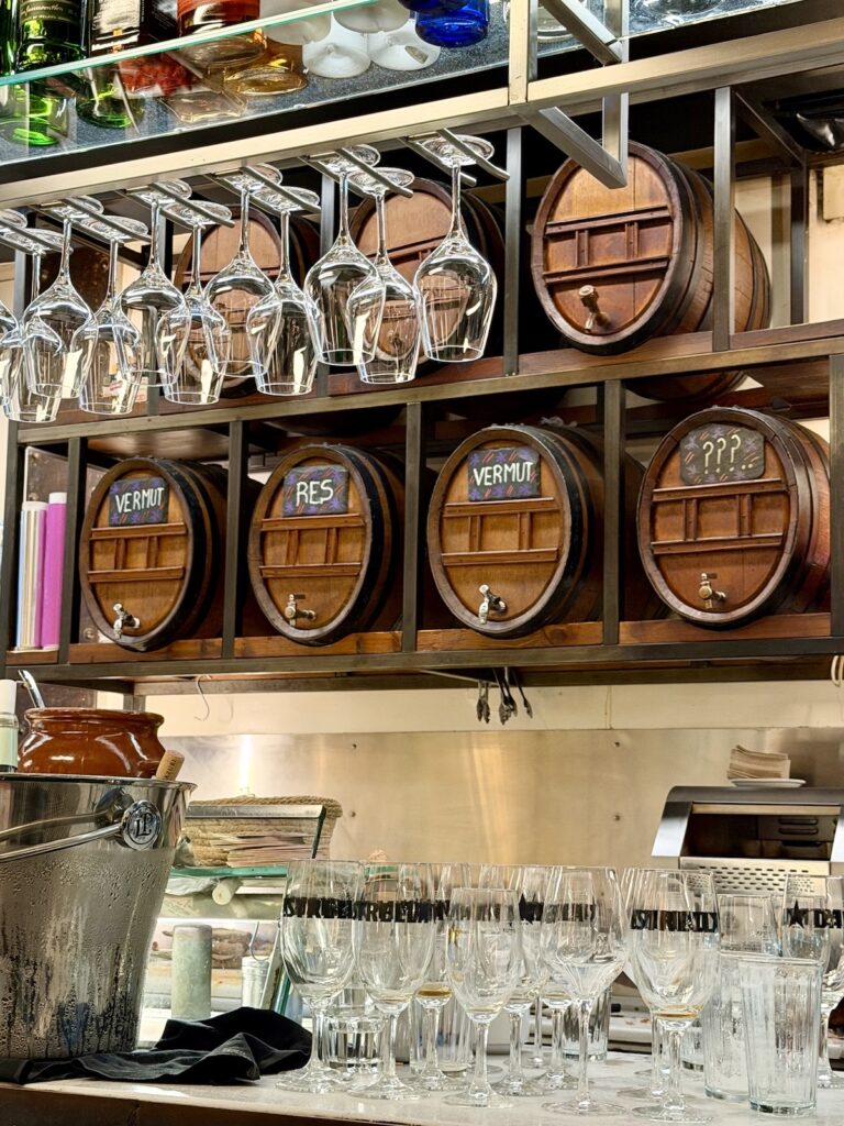 Innenansicht einer Weinbar in Barcelona mit traditionellen Holzfässern für Wermut und Wein, hängenden Weingläsern und einer gemütlichen, authentischen Atmosphäre.